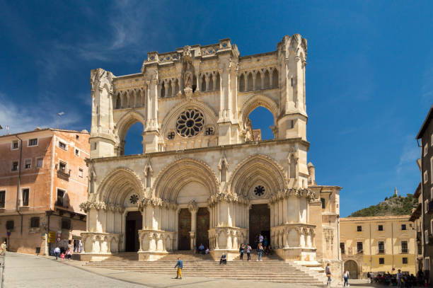 Facade of the Cathedral de Santa María la Mayor in Cuenca, Spain. Several people are passing by or sitting on the stairs. Sunny weather. 