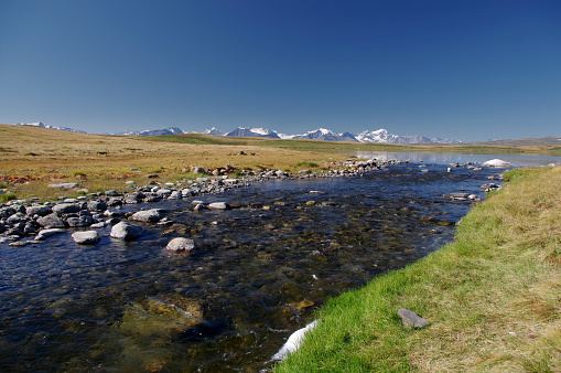 Highland river stream flowing from lake on a plateau on the background of mountains with snow and glaciers under clear blue sky Ukok, Altai mountains Siberia, Russia
