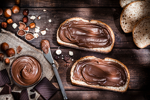 Top view of two toasts with chocolate and hazelnut spread shot on rustic wooden table. A glass bowl filled with chocolate spread and a knife are beside the toasts. Some shelled and peeled hazelnuts and chocolate pieces complete the composition. Predominant color is brown. Low key DSRL studio photo taken with Canon EOS 5D Mk II and Canon EF 100mm f/2.8L Macro IS USM