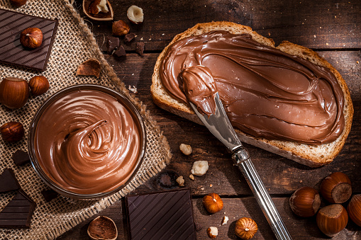 Top view of a toast with chocolate and hazelnut spread shot on rustic wooden table. A glass bowl filled with chocolate spread and a knife are beside the toast. Some shelled and peeled hazelnuts and chocolate pieces complete the composition. Predominant color is brown. Low key DSRL studio photo taken with Canon EOS 5D Mk II and Canon EF 100mm f/2.8L Macro IS USM