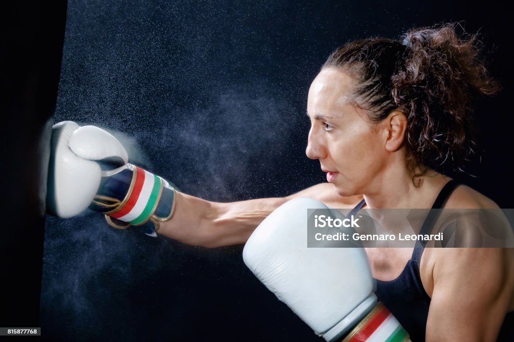 A boxer woman throws a fist. Fist in the sack. A boxer woman throws a punch in the sack. Splashes of water and sweat on the black background. Active Lifestyle Stock Photo