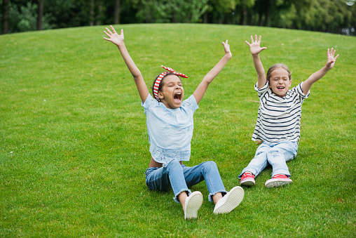 Cheerful multiethnic girls raising hands while sitting on green meadow