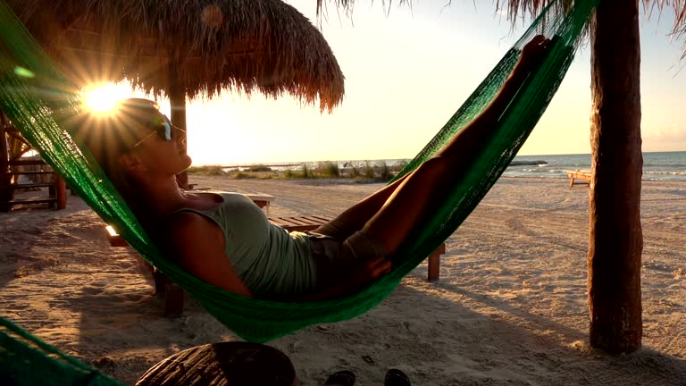 SLOW MOTION: Young woman swinging in hammock overlooking sandy beach at sunset