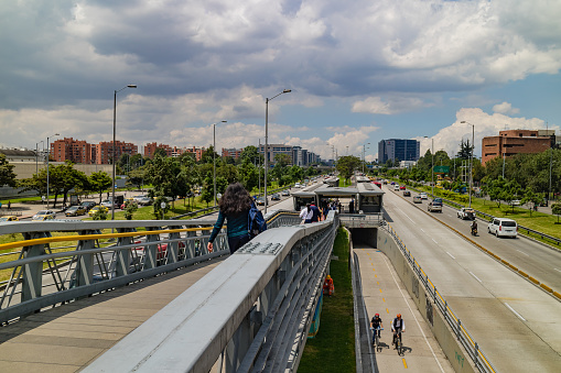 Bogotá, Colombia - February 16, 2017: Looking from the TransMilenio pedestrian walkway to the Salitre El Greco TransMilenio Station on Avenida El Dorado or Calle 26 and beyond; the road leads to Bogota's El Dorado International Airport. In the foreground is the walkway to the Station. It is lunchtime and the traffic on the Street is not as heavy as it normally can be. Also seen is the Cycle Lane that goes under the Station; a couple of cyclists are seen in the foreground. Photo shot in the early afternoon sunlight; horizontal format.