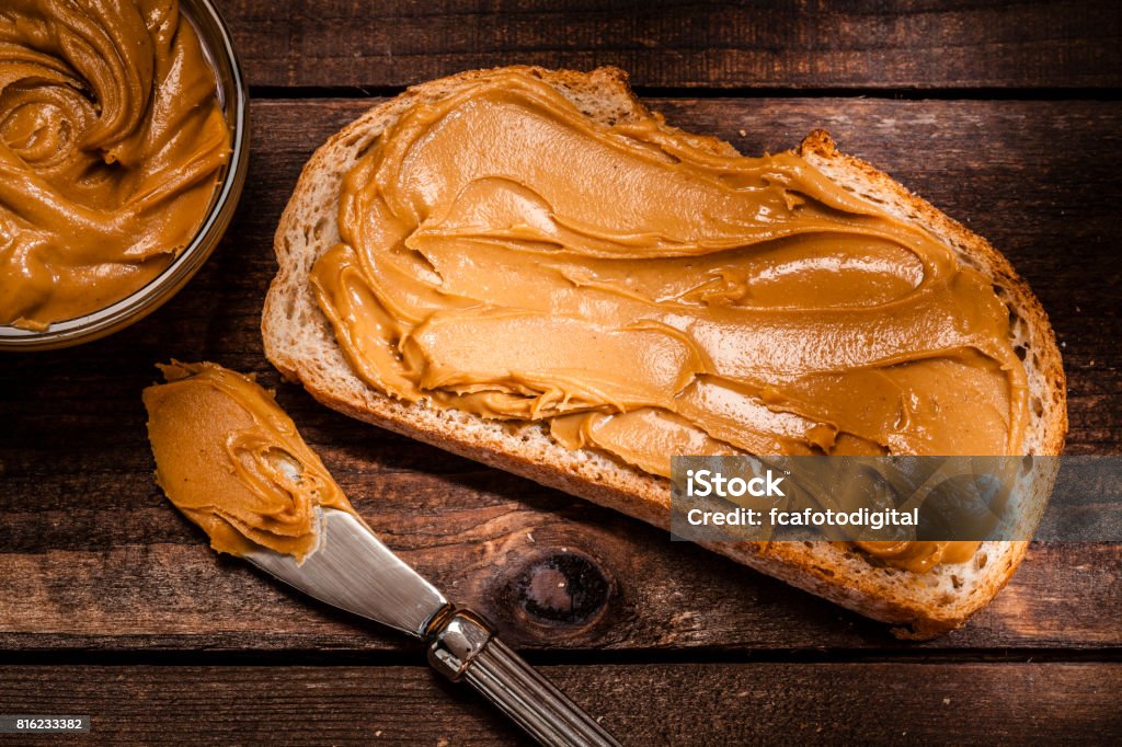 Peanut butter on bread slice shot on rustic wooden table Top view of two toasts with peanut butter shot on rustic wooden table. A little glass bowl filled with peanut butter and a knife is beside the toasts. Some shelled and peeled peanuts nuts complete the composition. Predominant color is brown. Low key DSRL studio photo taken with Canon EOS 5D Mk II and Canon EF 100mm f/2.8L Macro IS USM Peanut Butter Stock Photo