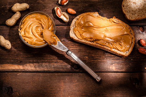 High angle view of a toast with peanut butter shot on rustic wooden table. A little glass bowl filled with peanut butter and a knife are beside the toast. Some shelled and peeled peanuts nuts complete the composition. Predominant color is brown. Low key DSRL studio photo taken with Canon EOS 5D Mk II and Canon EF 100mm f/2.8L Macro IS USM
