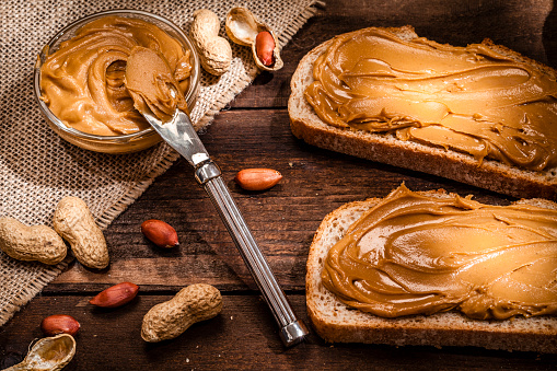 High angle view of two toasts with peanut butter shot on rustic wooden table. A little glass bowl filled with peanut butter and a knife are beside the toasts. Some shelled and peeled peanuts nuts complete the composition. Predominant color is brown. Low key DSRL studio photo taken with Canon EOS 5D Mk II and Canon EF 100mm f/2.8L Macro IS USM