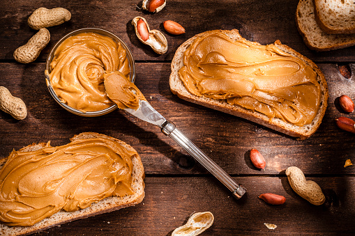 Top view of two toasts with peanut butter shot on rustic wooden table. A little glass bowl filled with peanut butter and a knife are between the toasts. Some shelled and peeled peanuts nuts complete the composition. Predominant color is brown. Low key DSRL studio photo taken with Canon EOS 5D Mk II and Canon EF 100mm f/2.8L Macro IS USM