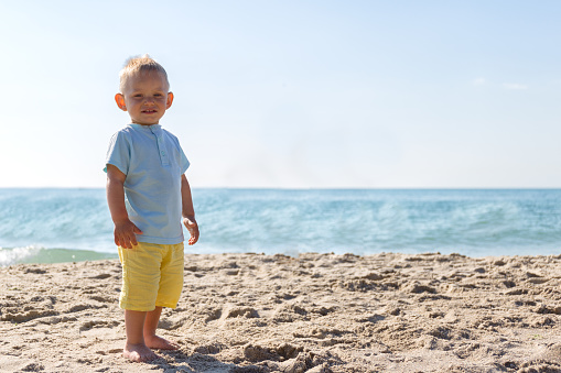 Summer,bright day,beach.Boy 1-2 years Caucasian blonde is on the sand and looking forward.The background of blue sea and sky.The concept of vacation,holidays