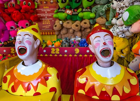 Two laughing clown ball machines at a fun fair with stuffed toys and other prizes in the background