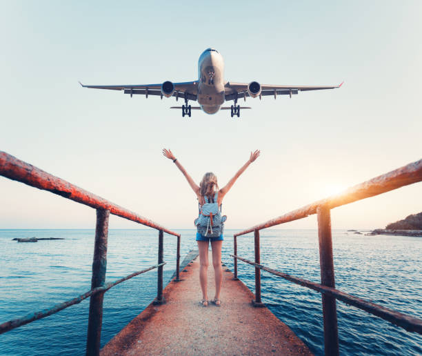 Airplane and woman at sunset. Summer landscape with girl standing on the sea pier with raised up arms and flying passenger airplane. Woman and landing commercial plane in the evening. Lifestyle Airplane and woman at sunset. Summer landscape with girl standing on the sea pier with raised up arms and flying passenger airplane. Woman and landing commercial plane in the evening. Lifestyle landing craft stock pictures, royalty-free photos & images