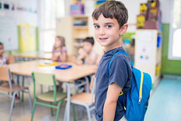 Cute schoolboy carrying backpack in classroom Side view portrait of cute schoolboy standing in classroom. Smiling male student is carrying backpack at school. He is wearing casuals. 6 7 years stock pictures, royalty-free photos & images
