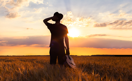 Person standing with his guitar in the warm summer fields looking at sunset for musical inspiration