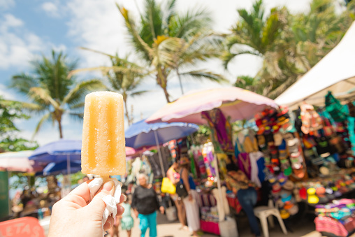 This is a horizontal color photograph of a yellow popsicle being held in a woman's hand on a hot day in the street market area of Sayulita, Mexico. Vendors are visible in the background.
