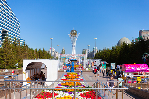 Astana, Kazakhstan - July 7, 2017: On a sunny day, people walk on the street and the square, parents and children rest on a beautiful day. Astana is a city of possibilities, very beautiful and tidy. In the foreground there are planted colorful flowers, and in the background of the building are huge and the Baiterek monument is a symbol of Astana. Blue sky without clouds.