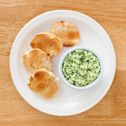 Top view of white dish with bread toasts and bowl with ricotta and pesto dip on it
