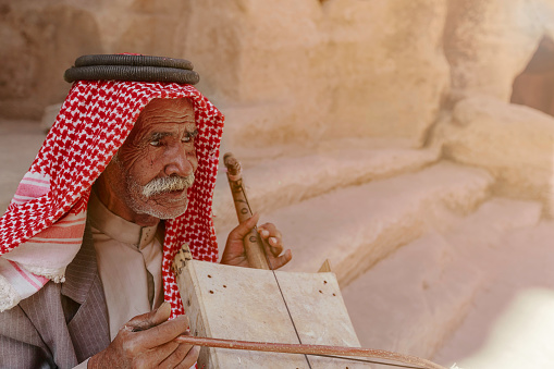 Little Petra, Jordan - June 20, 2017: Old Bedouin man or Arab man in traditional outfit, playing his musical instrument at the doorway of Little Petra.