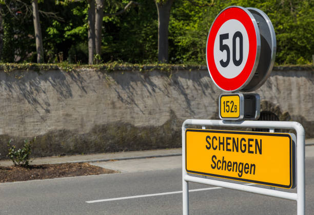 Road sign showing a speed limit of 50 and a yellow town sign for Schengen near a concrete wall and trees.