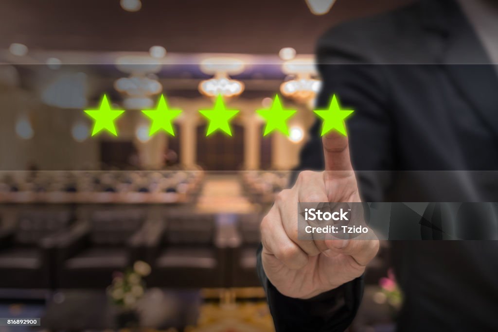 Businessman pointing five star symbol to increase rating of hotel over Abstract blurred photo of conference hall or seminar room with attendee background, business evaluation concept, Increase rating First Class Stock Photo