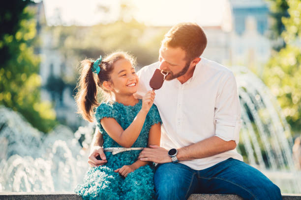 Young father and daughter eating ice-cream at the fountain Little girl in the park giving ice-cream to hеr father childrens day photos stock pictures, royalty-free photos & images