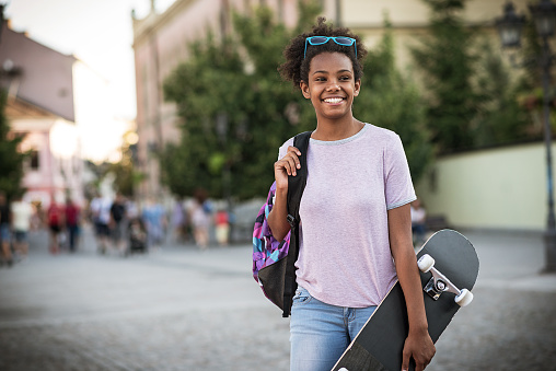 Summer is coming to an end and school children and high school students are getting ready for the responsibilities and tasks. Beautiful black girl with skateboard and backpack going to school.