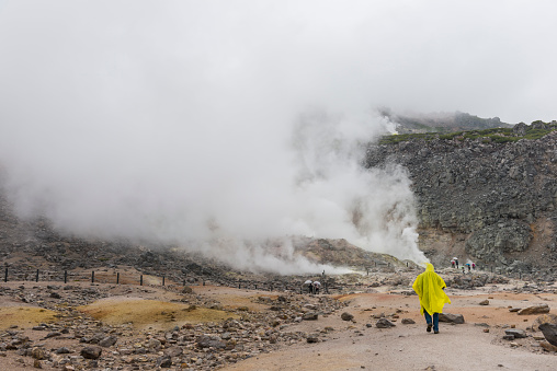 Front view a men in yellow raincoat walking to the sulfur area. In distance unrecognizable persons. Oyunuma pound in Noboribetsu, steaming pond, Hokkaido, Japan