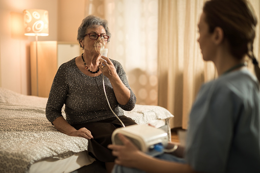 Senior woman having a visit from home caregiver and breathing through mask during medical oxygen therapy.