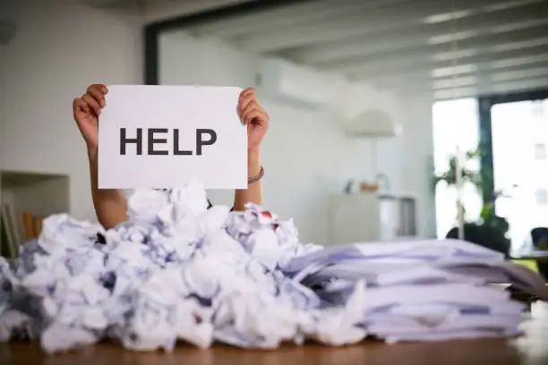 Shot of an unidentifiable businesswoman holding up a sign that reads "help" while drowning under a pile of paperwork in the office