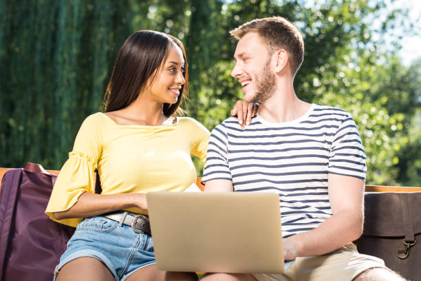 istock Happy multiethnic couple using laptop while sitting on bench in park and smiling each other 817327852