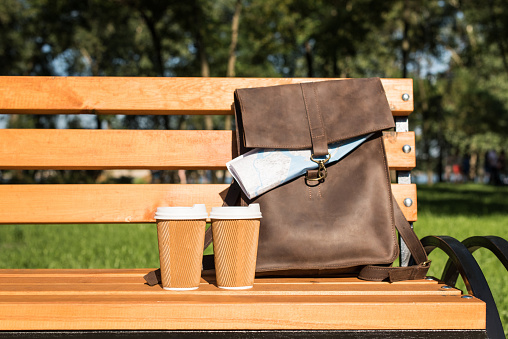 Close-up view of brown leather bag with map and disposable coffee cups on wooden bench