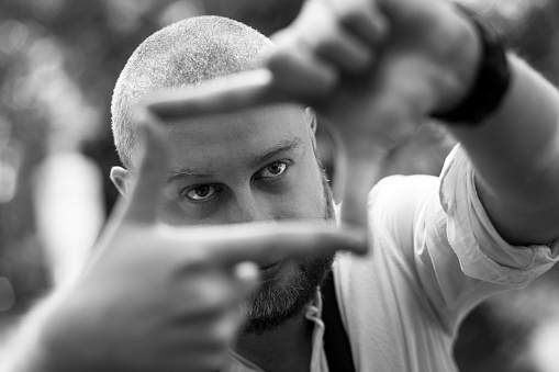 Close up image of a young male caucasian photographer framing a shot using his fingers. The focus is on his face which we can see through the frame he is making with his fingers. The man, in his 30s, has a shaved head and a beard. Outdoors summer image with green trees and foliage defocused in the background. Horizontal monochrome image with copy space.