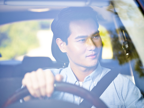 young asian man driving a vehicle, looking at scenery,  seen through the windshield glass.