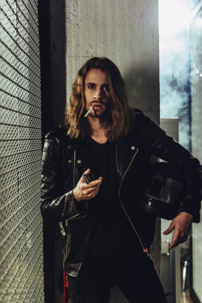 istock Handsome long haired man in leather jacket holding helmet and lighting cigarette with lighter 817347424