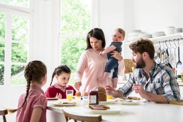 Family sitting at breakfast table, mother holding baby boy, father and girls eating food Two girls having breakfast with their dada as their mum carries their younger brother 6 11 months stock pictures, royalty-free photos & images