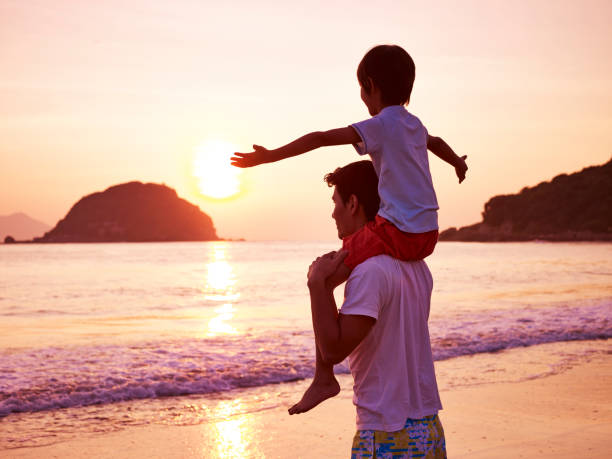 young asian father carrying son on shoulder on beach at sunrise.