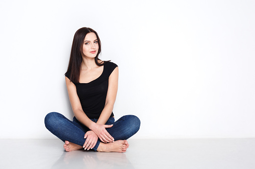 Home relax. Barefoot pensive woman in casual wear sitting indoors on floor by white blank wall, copy space