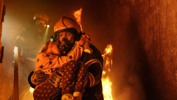 Brave Fireman Descends Stairs of a Burning Building and Holds Saved Girl in His Arms. Open fire and one Firefighter in the Background. Brave Fireman Descends Stairs of a Burning Building and Holds Saved Girl in His Arms. Open fire and one Firefighter in the Background. rescue stock pictures, royalty-free photos & images