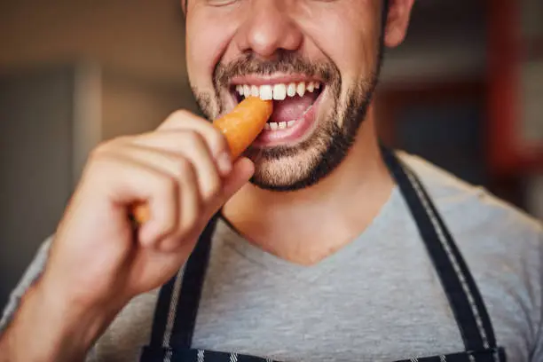 Cropped shot of an unidentifiable young man eating a carrot in his kitchen at home