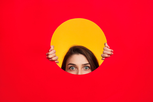 Crop woman looking out of circle cut in red paper on yellow background.