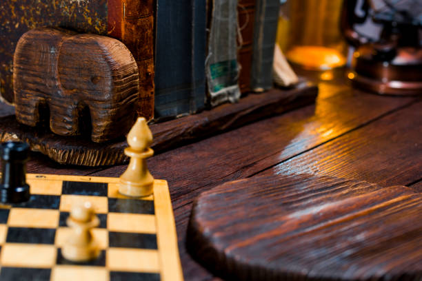 Private library desk of mahogany. Vintage books on shelf with wooden holder in the shape of elephant. Chess board with pieces and serving board on the desk. Close-up. Copy space
