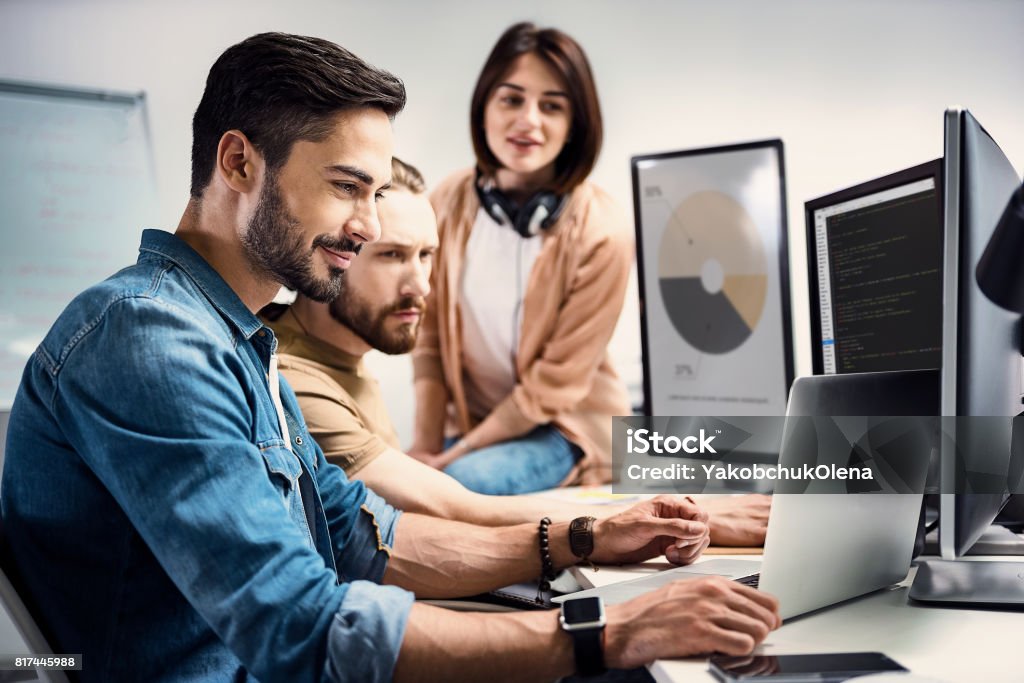 Cheerful man demonstrating information to partners Side view smiling bearded male worker showing data on laptop to colleagues while sitting at table in modern apartment Teamwork Stock Photo