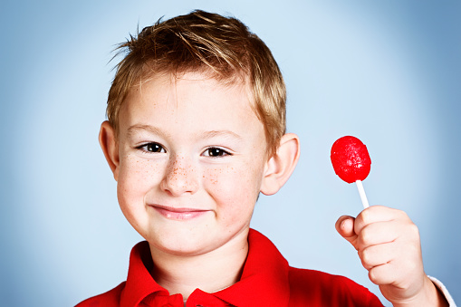 A cute, freckled pre-school boy grins mischievously as he holds up a red lollipop sweet. Delicious!