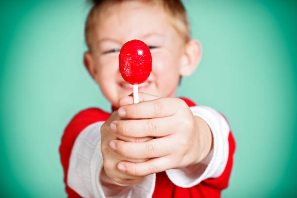 A smiling pre-school boy holds his red lolly out towards camera, offering a taste of his sweet treat.