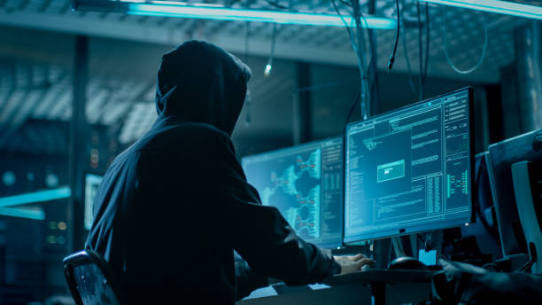 Shot from the Back to Hooded Hacker Breaking into Corporate Data Servers from His Underground Hideout. Place Has Dark Atmosphere, Multiple Displays, Cables Everywhere. Shot from the Back to Hooded Hacker Breaking into Corporate Data Servers from His Underground Hideout. Place Has Dark Atmosphere, Multiple Displays, Cables Everywhere. hacker stock pictures, royalty-free photos & images