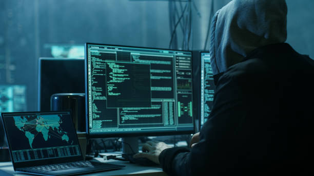istock Dangerous Hooded Hacker Breaks into Government Data Servers and Infects Their System with a Virus. His Hideout Place has Dark Atmosphere, Multiple Displays, Cables Everywhere. 817486220