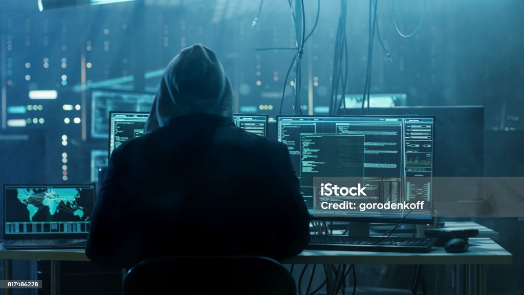 Dangerous Hooded Hacker Breaks into Government Data Servers and Infects Their System with a Virus. His Hideout Place has Dark Atmosphere, Multiple Displays, Cables Everywhere. Computer Hacker Stock Photo