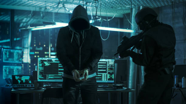 istock Handcuffed Masked Hacker is Standing and Guarded by Fully Armed Special Forces Soldier. They're in Hacker's Hideout Basement with Multiple Operating Displays. 817486294