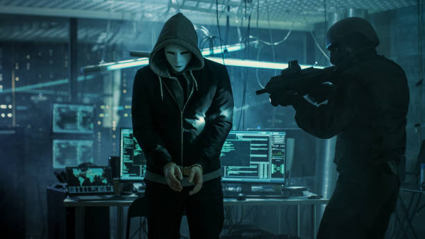 istock Masked Hacker Wearing Handcuffs is Guarded by Fully Armed Special Forces Soldier.  They're in Hacker's Hideout Basement with Multiple Operating Displays. 817486330