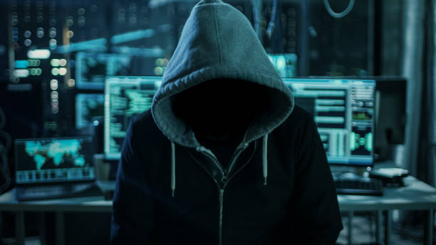 istock Dangerous Internationally Wanted Hacker Wth Hided Face Looking into the Camera. In the Background His Dark Operating Room with Multiple Displays and Cables. 817486350