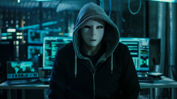 istock Dangerous Internationally Wanted Hacker with Covered Face Looking into the Camera. In the Background His Operating Room with Multiple Displays and Cables. 817486370